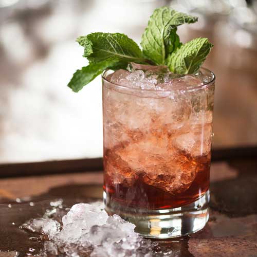 Short transparent glass on wooden surface with red colored liquid cocktail inside - the Old Forester Hibscus Julep.